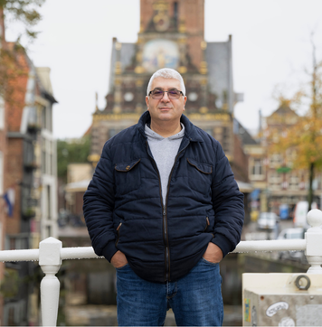 Kandidaat staat op brug met historisch gebouw op de achtergrond.