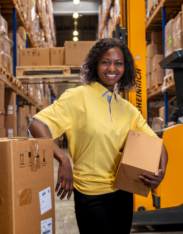 Inpakmedewerker in gele polo houdt dozen vast in een groot magazijn en glimlacht naar de camera.
