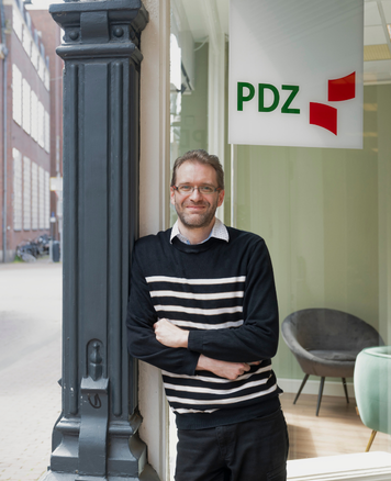 Kandidaat leunt naast een zuil bij de ingang van een PDZ-kantoor met zichtbaar bedrijfslogo.