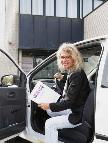 Zorgmedewerker zit in een bedrijfsauto met een map in de hand voor een gebouw van een thuiszorgorganisatie.