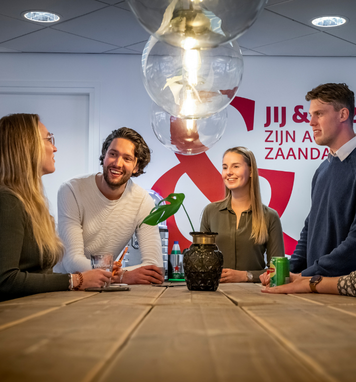 Vier collega’s staan lachend bij elkaar aan een tafel in een modern kantoor tijdens een informeel overlegmoment.