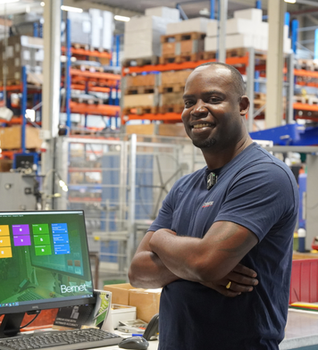 Logistiek medewerker staat in een magazijn bij een werkplek met computer, met op de achtergrond stellingen vol goederen.