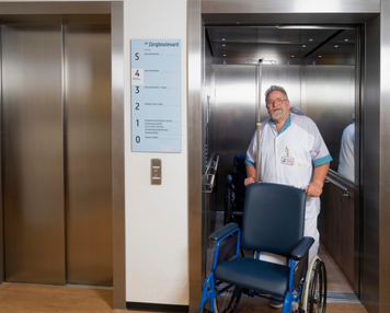 Persoon in zorgkleding staat met een rolstoel bij een geopende lift in een zorglocatie.