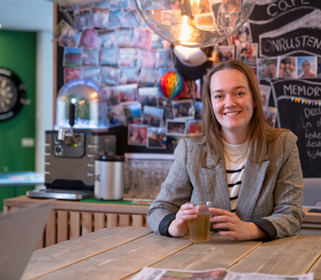 Persoon zit aan een tafel met een drankje in een gemeenschappelijke ruimte met koffiecorner en fotowand op de achtergrond.