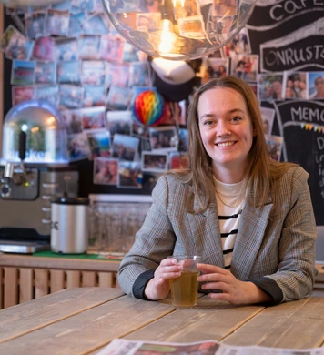 Persoon zit aan een tafel met een drankje in een gemeenschappelijke ruimte met koffiecorner en fotowand op de achtergrond.