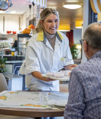 Helpende Plus serveert lachend een maaltijd aan een bewoner in een eetruimte van de zorginstelling.