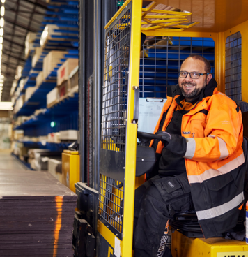 Reachtruckchauffeur in oranje werkjas lacht terwijl hij werkt in een houtmagazijn.