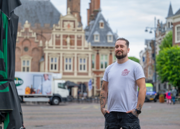 Kandidaat poseert zelfverzekerd op stadsplein met historisch gebouw op de achtergrond.