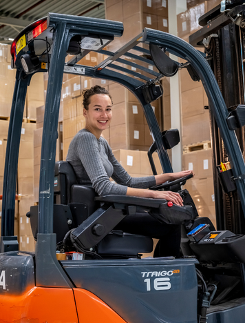 Heftruckchauffeur lacht terwijl ze in een magazijn vol dozen een heftruck bestuurt.