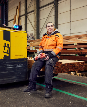 Logistiek medewerker zit in een loods op een stapel hout naast een heftruck en houdt een gereedschap vast.