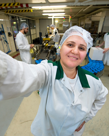 Productiemedewerker in witte werkjas en haarnet maakt een selfie in een productieomgeving met lopende band en collega’s op de achtergrond.