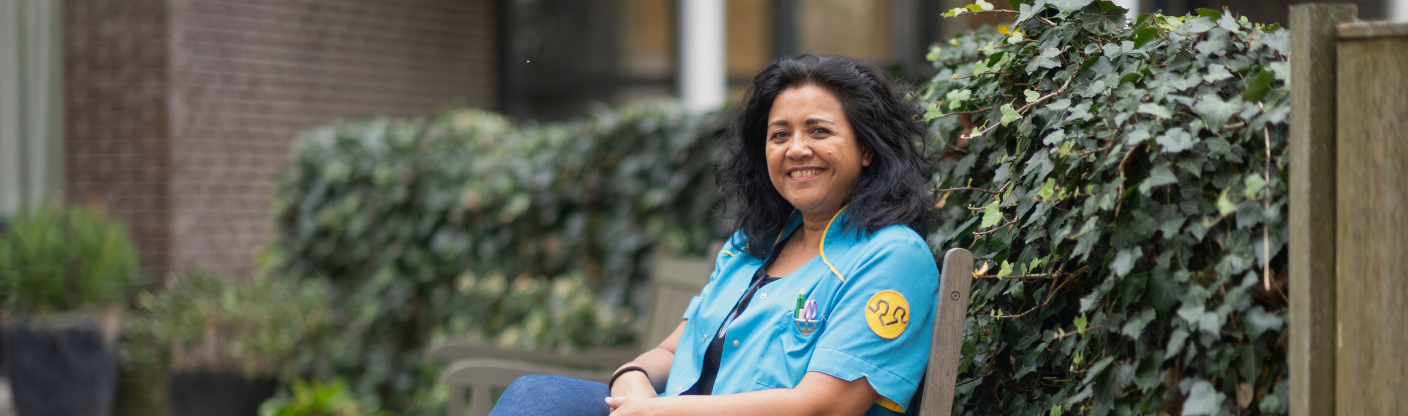 Zorgmedewerker in een blauw werkuniform zit op een houten bankje naast een groene haag op een buitenlocatie.