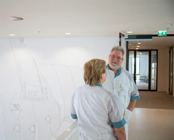 Twee medewerkers in werkkleding staan tegenover elkaar in een gang van een zorglocatie.