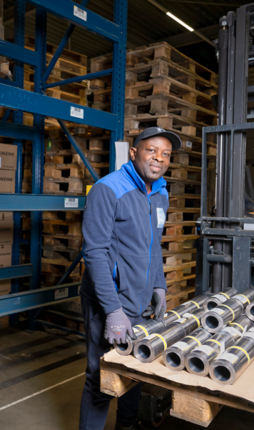 Heftruckchauffeur in magazijn lacht terwijl hij werkt met metalen rollen en pallets.