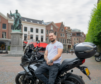 Kandidaat poseert met motor op stadsplein bij standbeeld.