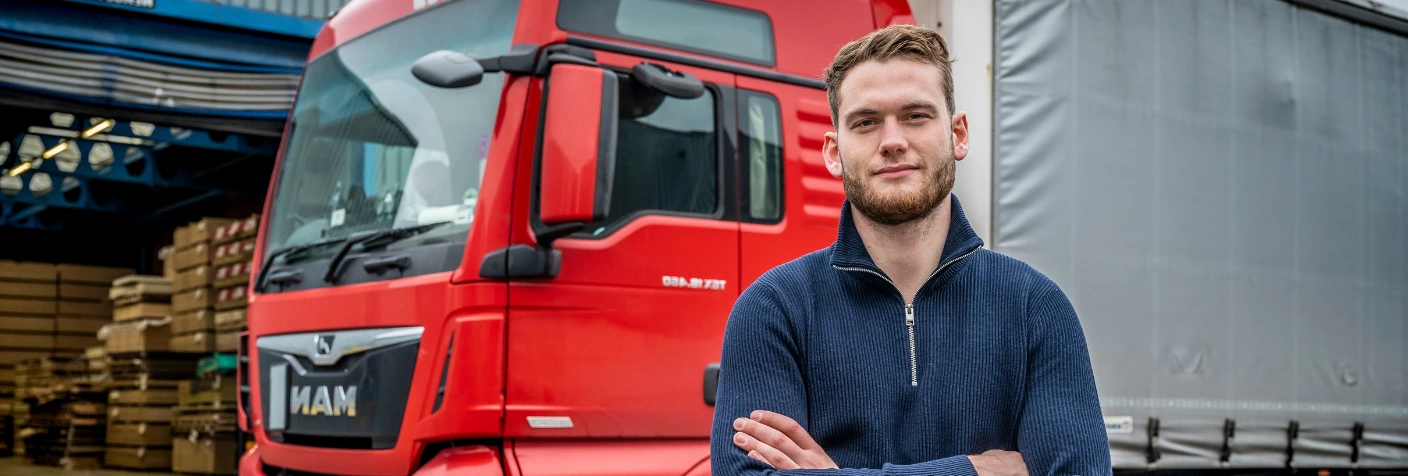 Transportmedewerker poseert voor rode vrachtwagen op bedrijventerrein.