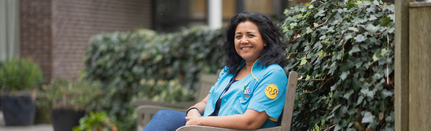 Verzorgende IG zit buiten op een bankje en glimlacht richting de camera.