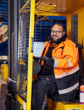 Reachtruckchauffeur in oranje werkjas lacht terwijl hij werkt in een houtmagazijn.