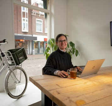 Kandidaat werkt aan een laptop aan een houten tafel in een kantoorruimte met een fiets en straatzicht op de achtergrond.