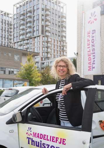 Zorgmedewerker staat naast een bedrijfsauto van Madeliefje Thuiszorg voor een zorglocatie.