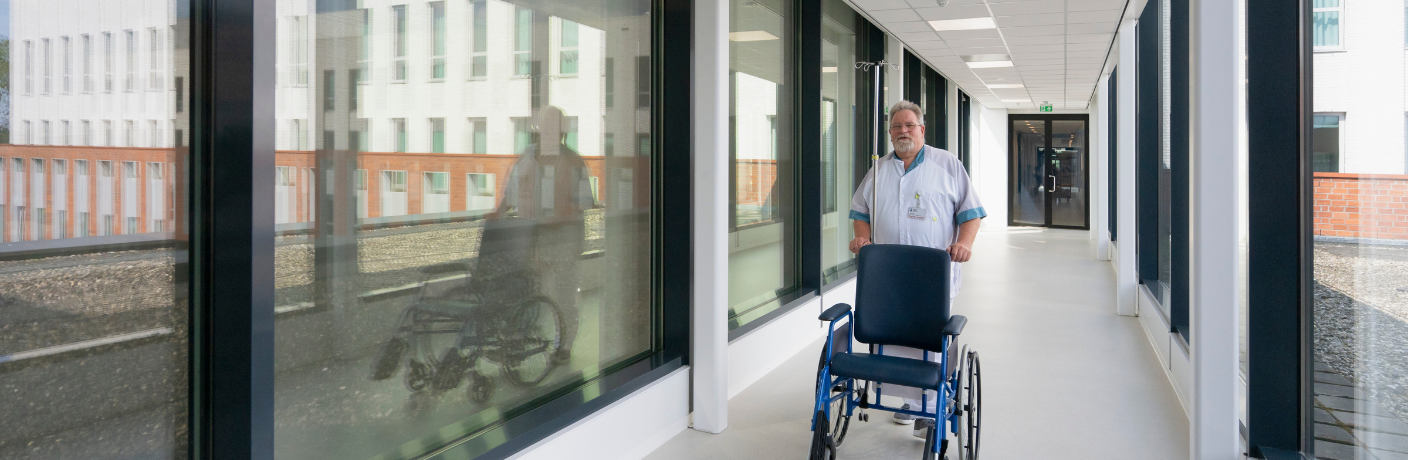 Persoon in zorgkleding duwt een rolstoel door een gang in een zorginstelling.