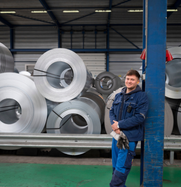 Operator in blauwe werkkleding poseert glimlachend in een fabriekshal met grote metalen rollen op de achtergrond.