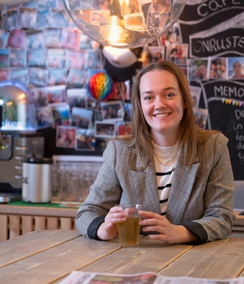 Persoon zit aan een tafel met een drankje in een gemeenschappelijke ruimte met koffiecorner en fotowand op de achtergrond.