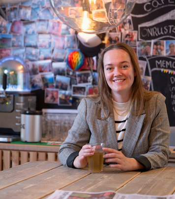 Persoon zit aan een houten tafel met een glas drinken in een kantoorruimte met fotowand en koffiemachine op de achtergrond.
