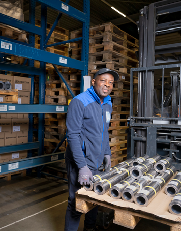 Heftruckchauffeur in magazijn lacht terwijl hij werkt met metalen rollen en pallets.