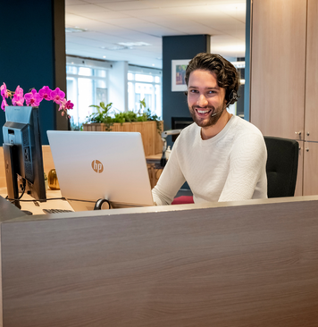 Receptionist met headset werkt achter bureau en glimlacht naar de camera.