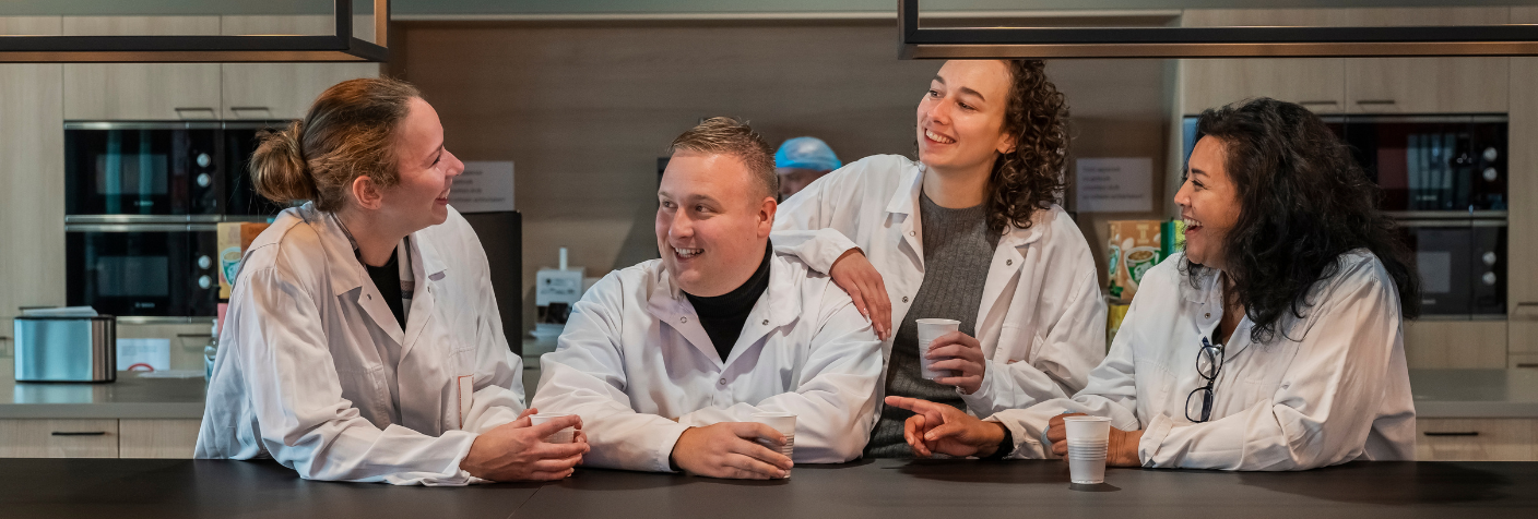 Vier productiemedewerkers in witte jassen zitten samen aan een tafel in een kantine, lachend en in gesprek tijdens een pauzemoment.