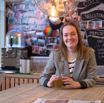 Persoon zit aan een houten tafel met een glas drinken in een kantoorruimte met fotowand en koffiemachine op de achtergrond.