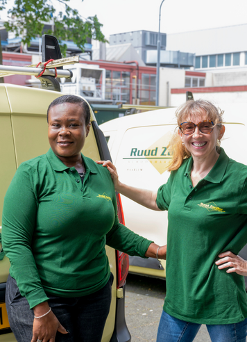 Twee kandidaten in groene werkkleding staan glimlachend naast een bedrijfswagen.