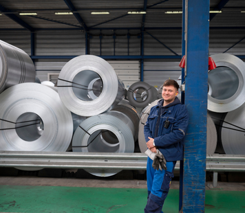 Operator in blauwe werkkleding poseert glimlachend in een fabriekshal met grote metalen rollen op de achtergrond.