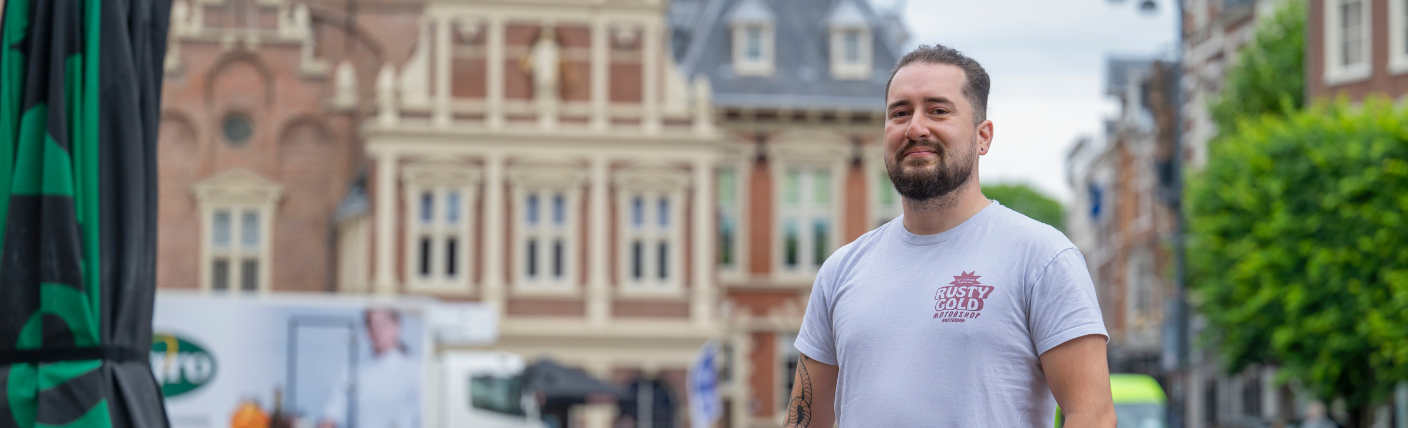 Kandidaat die ontspannen op straat poseert voor historische stadsgebouwen.