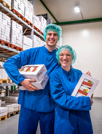 Twee inpakmedewerkers in blauwe werkkleding en haarnetjes houden dozen met verpakte producten vast in een magazijn.