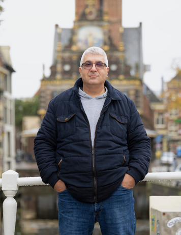 Kandidaat staat op brug met historisch gebouw op de achtergrond.