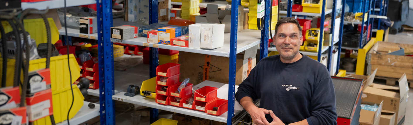 Monteur staat glimlachend in een magazijn tussen stellingen vol onderdelen en materiaal.