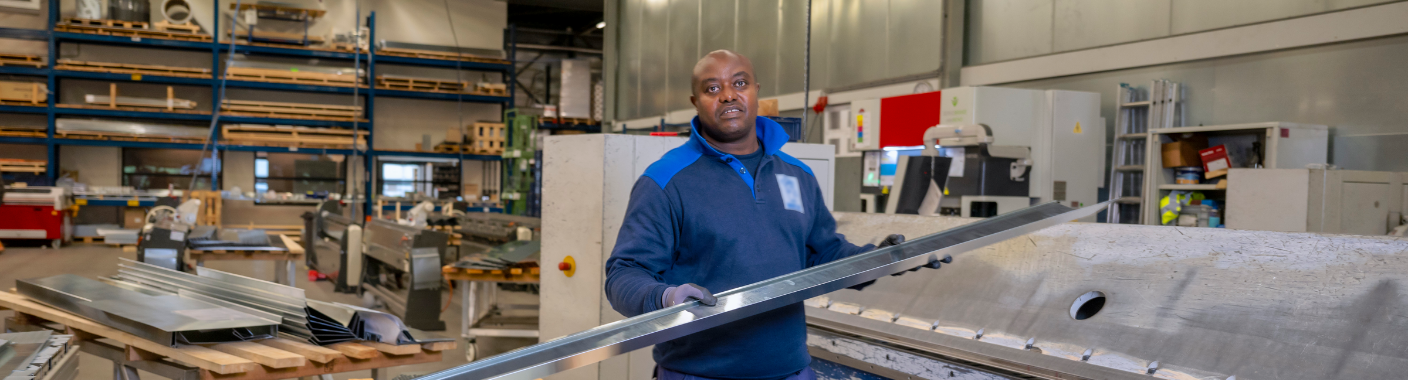 Technisch medewerker werkt met lange metalen profielen aan een machine in een industriële werkplaats.
