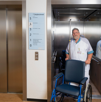 Verpleegkundige met rolstoel komt uit de lift van een ziekenhuis.