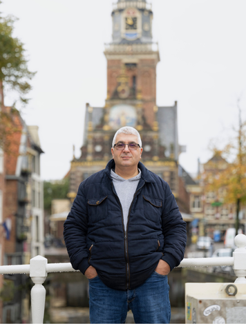 Kandidaat staat op brug met historisch gebouw op de achtergrond.