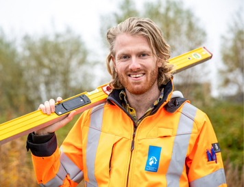 Persoon in een oranje veiligheidsjas houdt een waterpas op de schouder op een buitenlocatie.