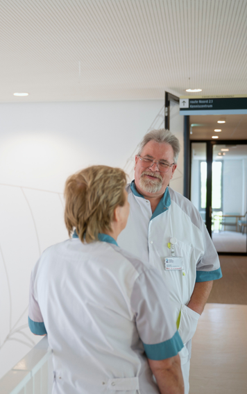 Twee zorgmedewerkers in werkkleding staan met elkaar in gesprek in een gang van een zorginstelling.