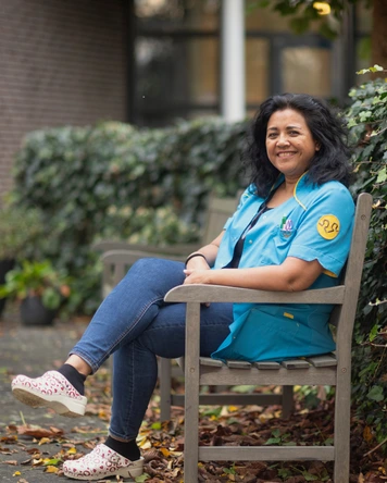 Zorgmedewerker zit op een houten bank buiten bij een gebouw met groenvoorziening op de achtergrond.