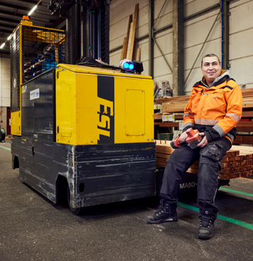 Heftruckchauffeur in oranje werkjas zit lachend op heftruck in houtmagazijn.