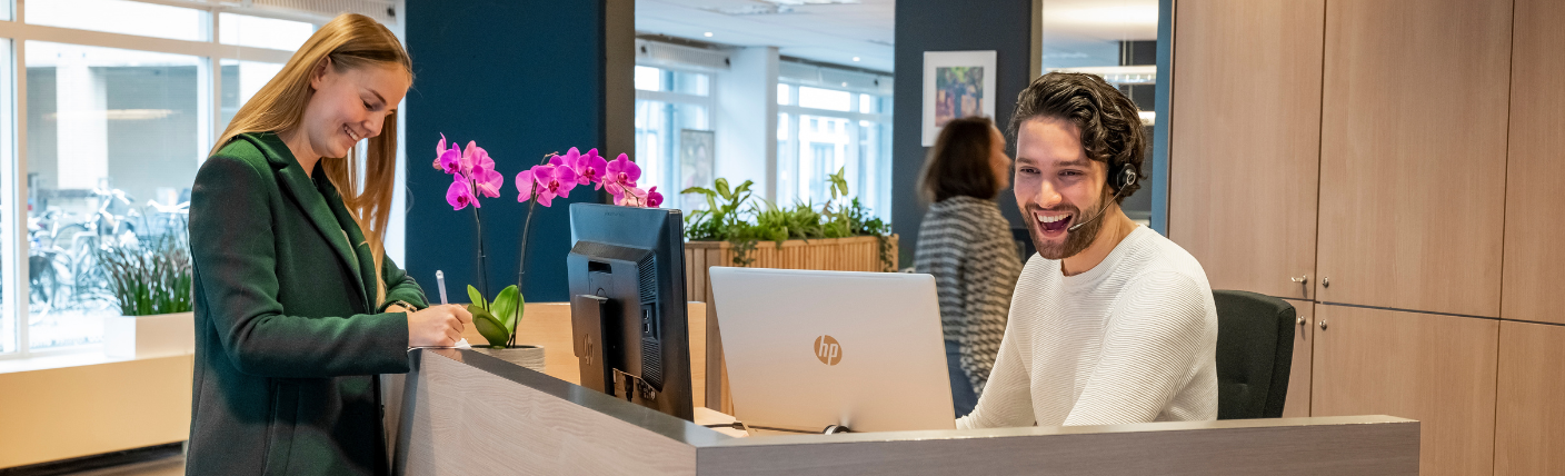 Receptionist helpt bezoeker aan de balie met een glimlach.