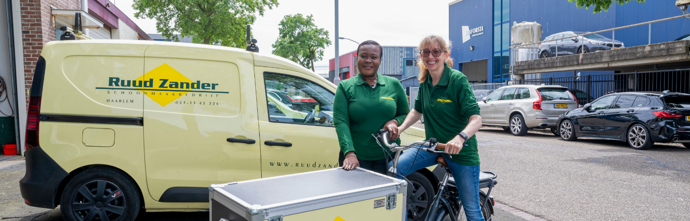 Twee schoonmaakmedewerkers in werkkleding staan bij een bakfiets naast een bedrijfsbus op een bedrijventerrein.