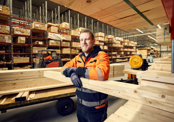 Persoon in veiligheidskleding staat bij houten balken in een magazijn met stellingen vol bouwmaterialen.