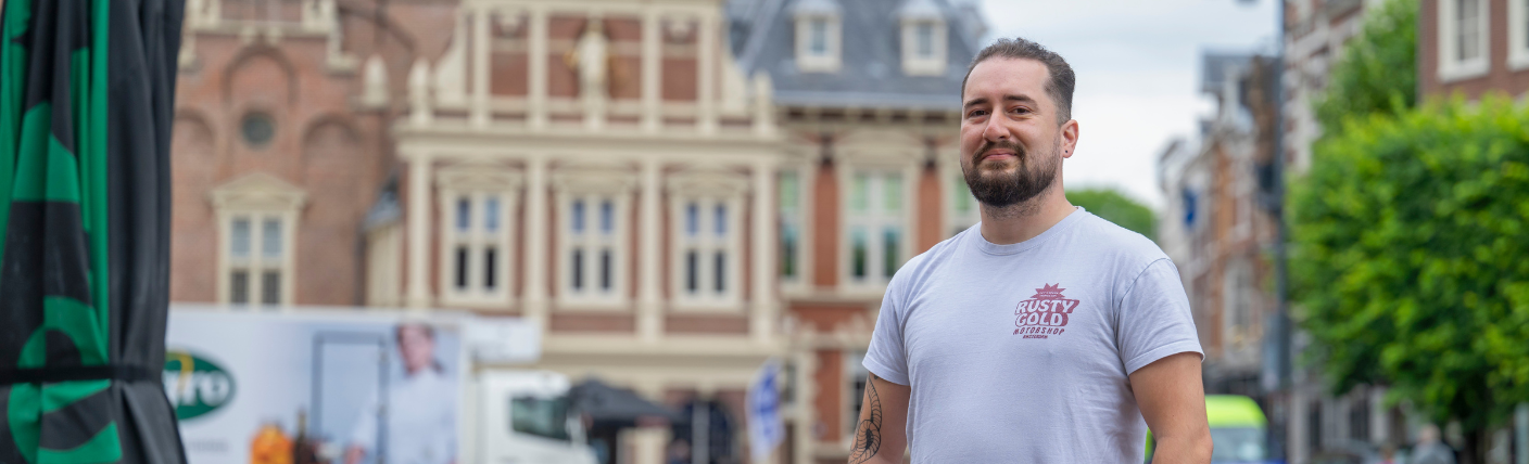 Kandidaat poseert op stadsplein met historisch gebouw op de achtergrond.