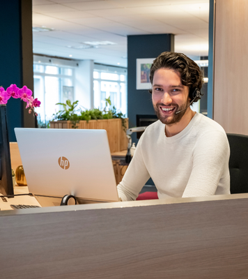 Administratief medewerker met headset werkt achter bureau en glimlacht naar de camera.